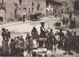 Vita di strada. Persone di ogni età, dai più piccoli agli anziani, assistono alla spettacolo di strada. Si notano botteghe, ambulanti, artigiani.